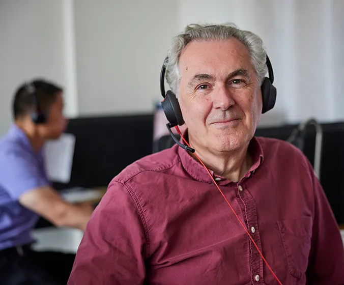 Peter som är volontär på Mind i kontorsmiljö. Han har ett headset på sig och ser rakt in i kameran ett lugnt leende.