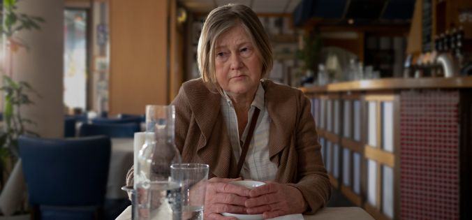 Äldre kvinna sitter ensam vid ett cafébord med händerna runt en kopp och en trött, tom blick.