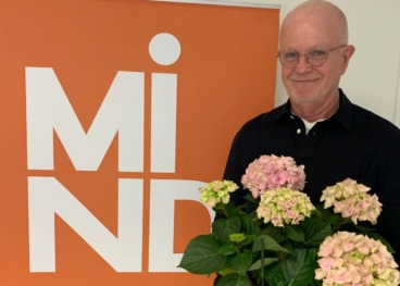 Lasse, volontär hos Mind, håller en bukett blommor och ler försiktigt mot kameran. I bakgrunden syns en rollup med Minds logotyp.
