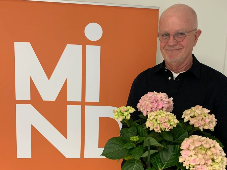 Lasse, volontär hos Mind, håller en bukett blommor och ler försiktigt mot kameran. I bakgrunden syns en rollup med Minds logotyp.