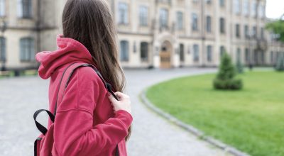 Skolstart. En ung kvinna i röd huvtröja blickar mot en skolbyggnad.