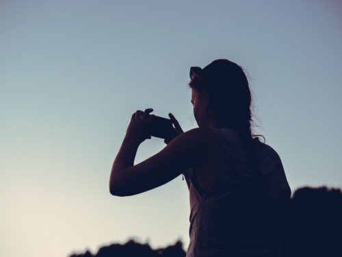 Ung kvinna i motljus fotograferar med sin telefon. Blå kvällshimmel.